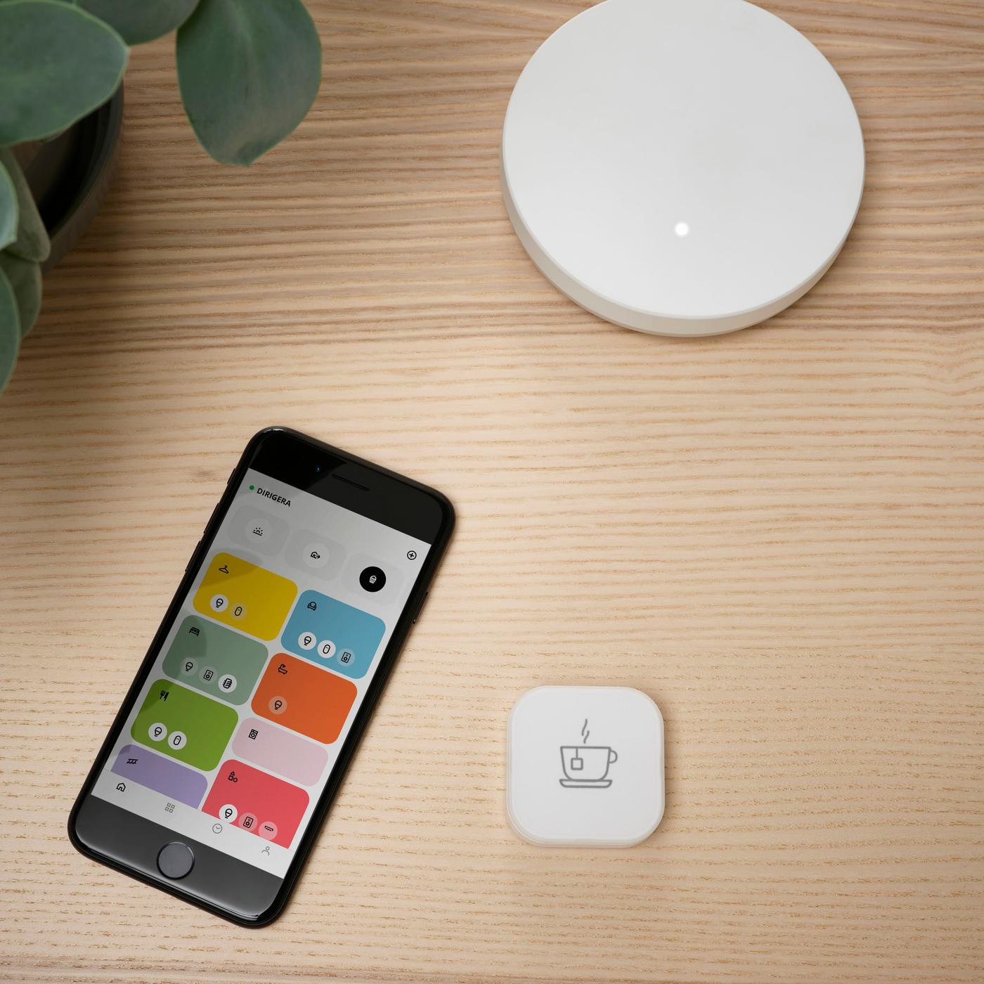 A phone displaying the IKEA Home smart app lying next to a DIRIGERA smart hub and a smart button.