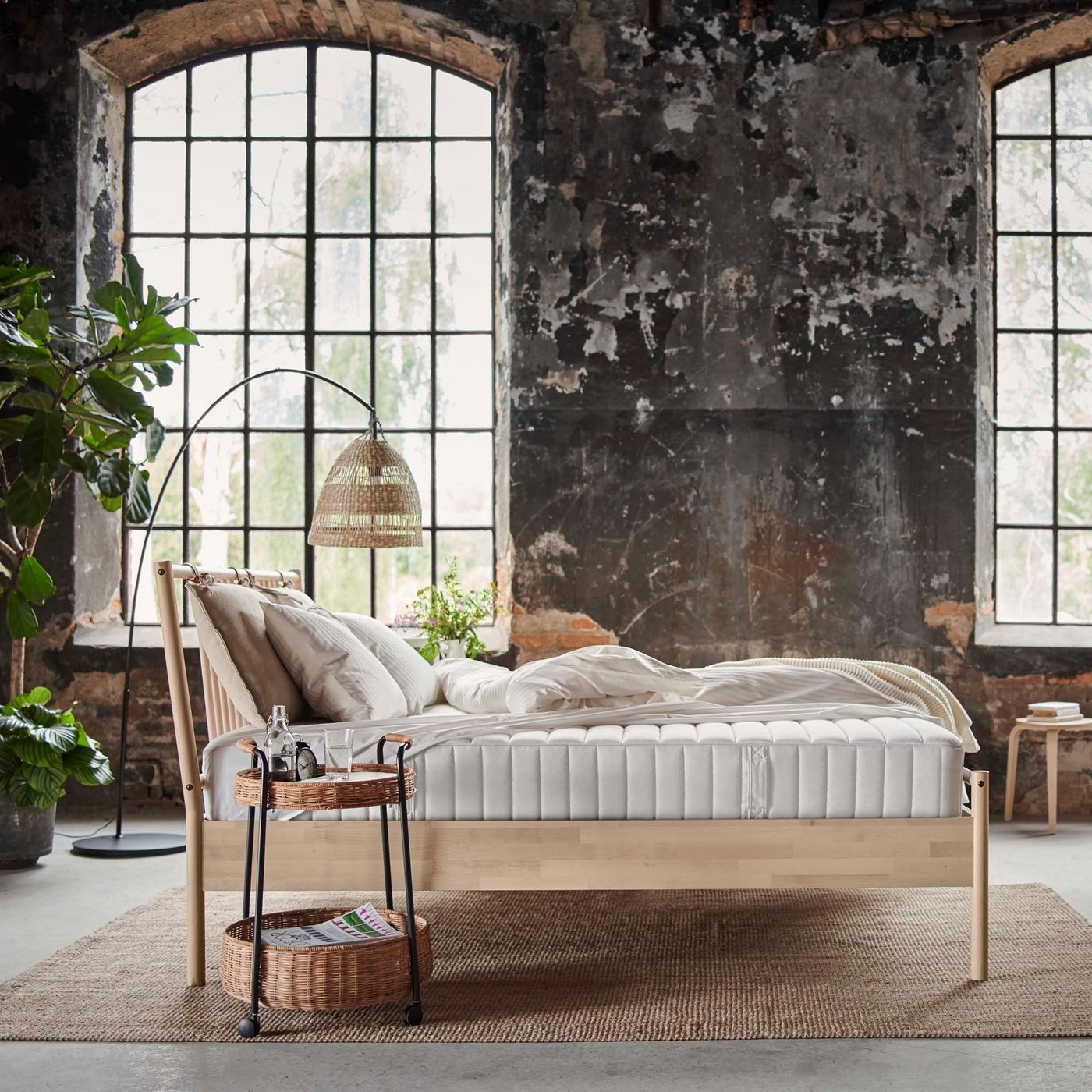 A bed with a VALEVÅG pocket sprung mattress in a room with a rattan bedside table and lamp nearby. In the background are two large windows and lots of lovely hanging plants.