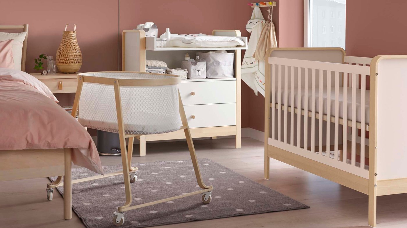 A basinett, cot and changing table stand in a pink and cream bedroom.