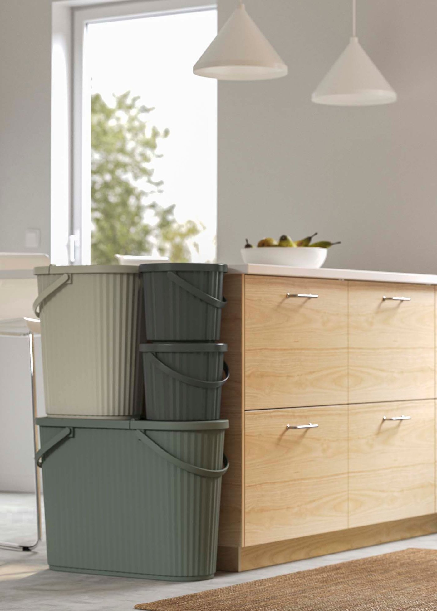 Three recycling bins in different shades of green are stacked against a wood kitchen island in a white kitchen.