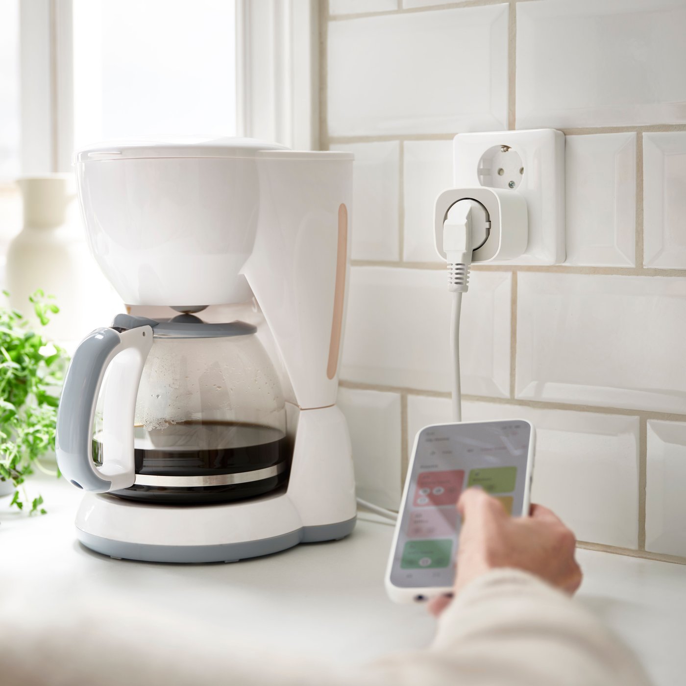 A hand is seen using the IKEA Home smart app. In the background, a coffee maker is plugged into INSPELNING - a smart plug that measures energy use.