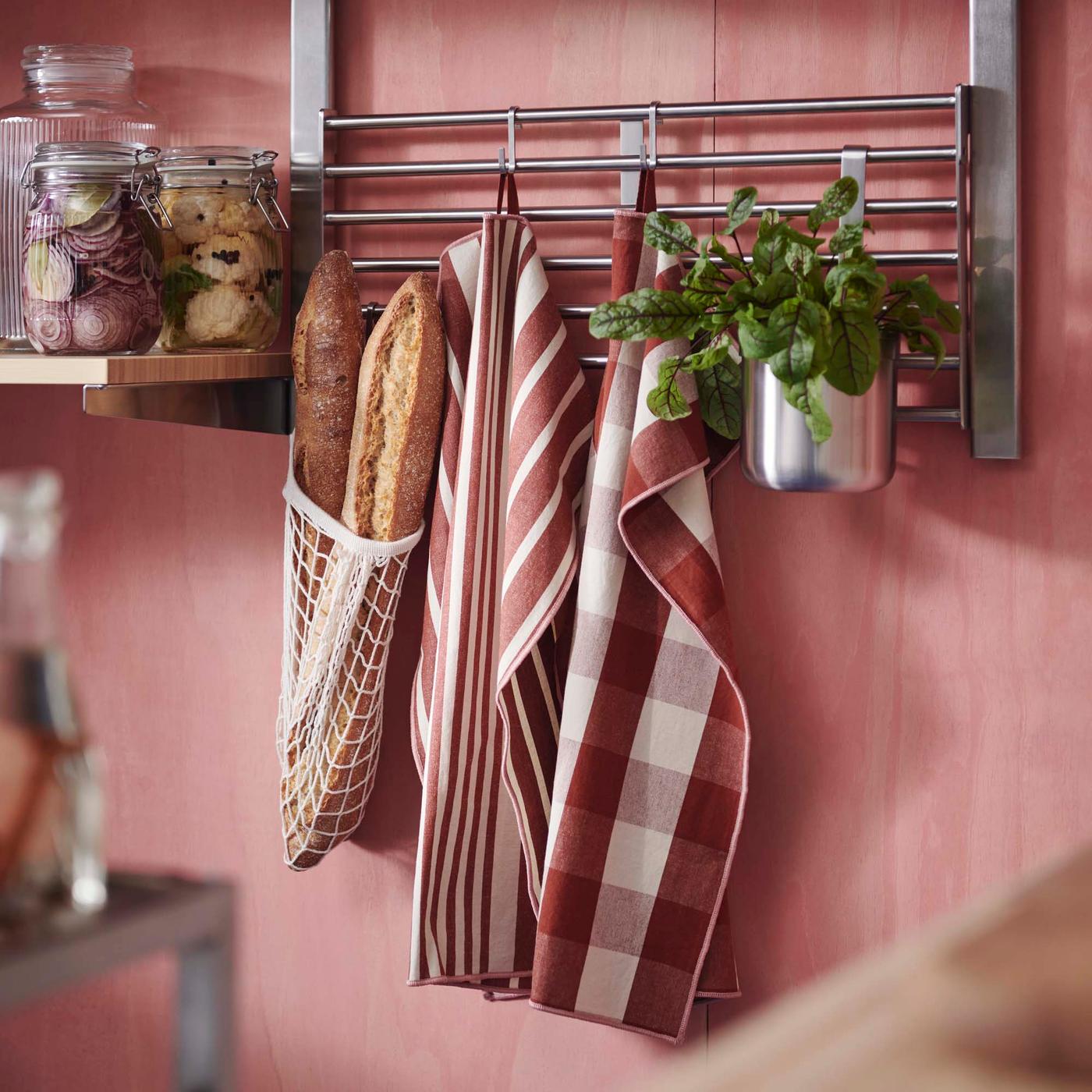 Checked tea-towels, herbs and a baguette in a white string shopping bag hang from a stainless-steel rack against a pink kitchen wall.