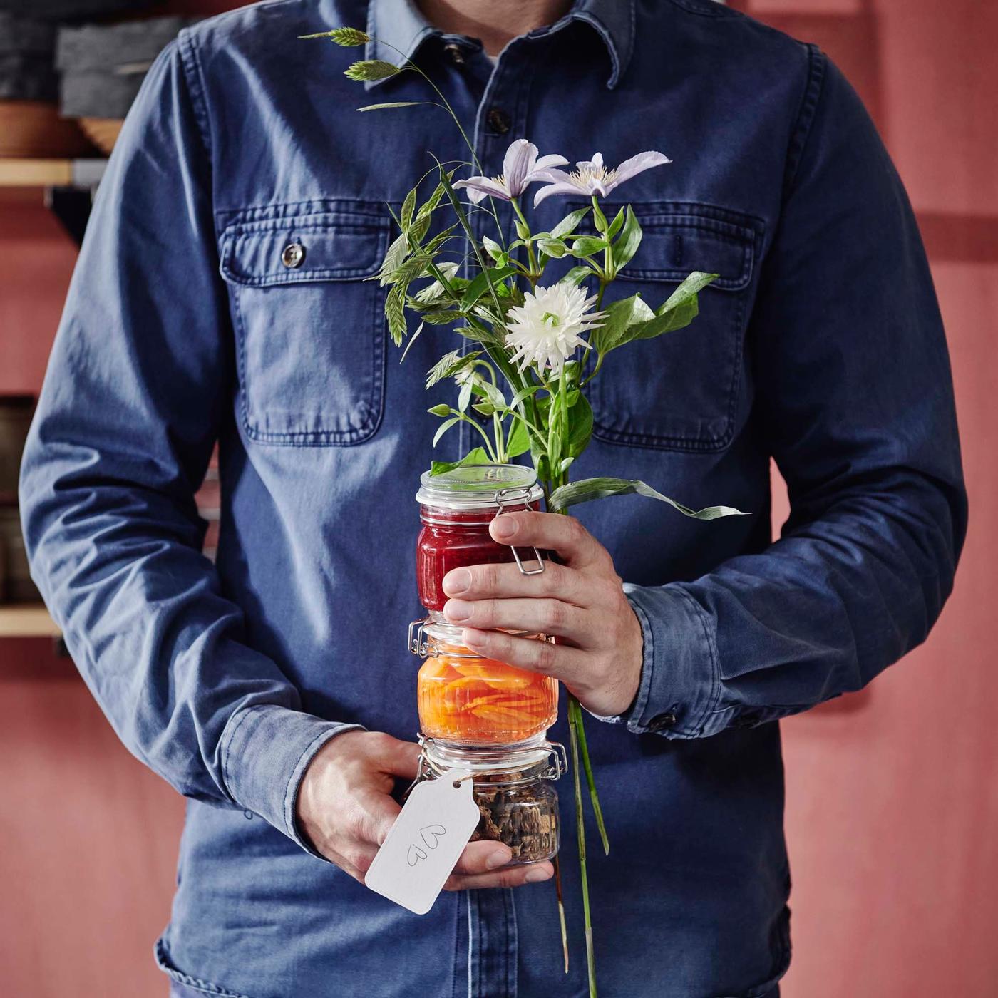 A person in a denim shirt holds a stack of glass jars with presserves in them, along with a bunch of white wildflowers.