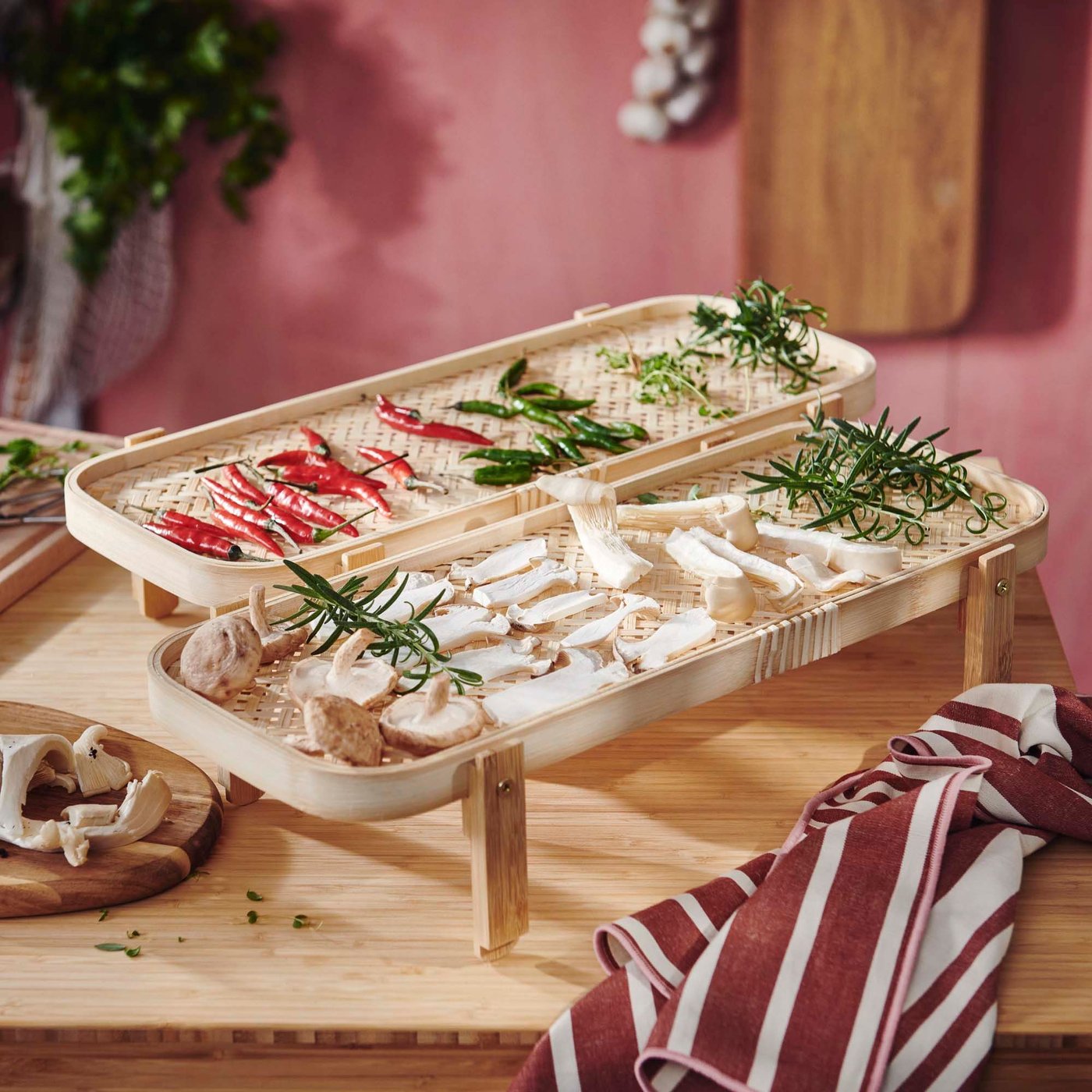 Herbs, mushrooms and chillies are spread out on two bamboo drying racks, side by side on a wood table.