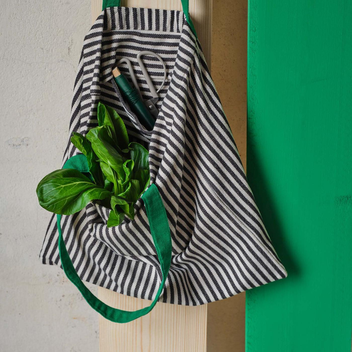 A black and white striped tote bag with green handles, with vegetables inside, hangs from a wall.