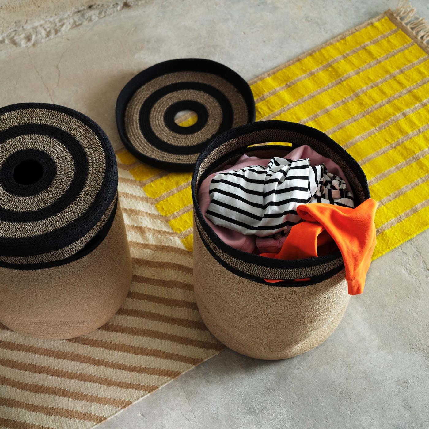 Two woven baskets and a woven plate, all from the MÄVINN collection, on a yellow rug.