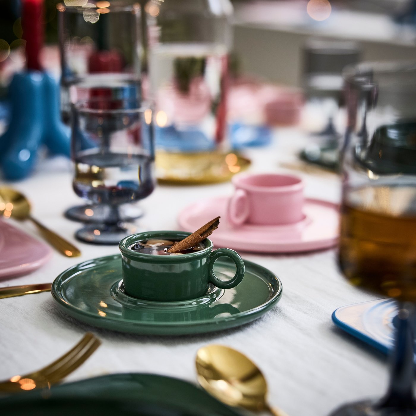 A green cup with saucer holding mulled wine and a vanilla stick, with glasses and a matching pink cup in the background.