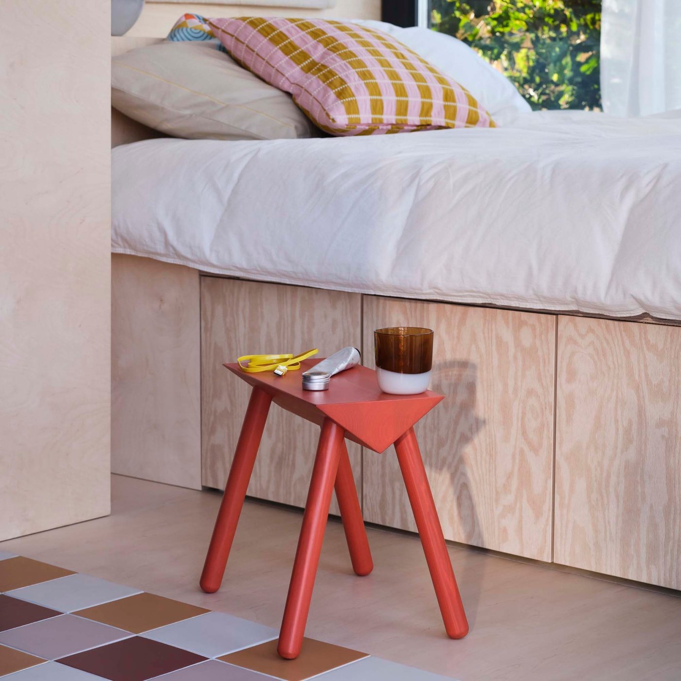 A red-orange OMMJÄNGE stool with a glass and objects on top, next to a wood base bed and a chequerboard floor.