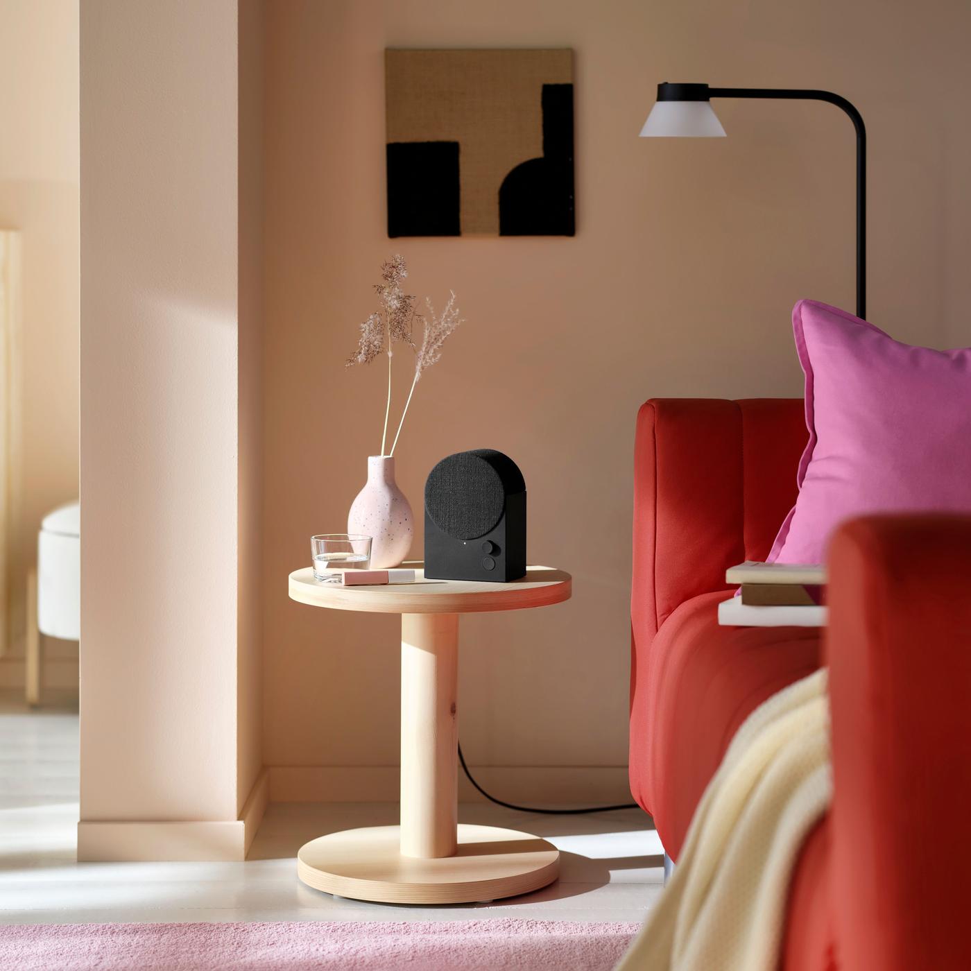 A black speaker on a wooden sideboard beside a pink vase and a red sofa in a room.