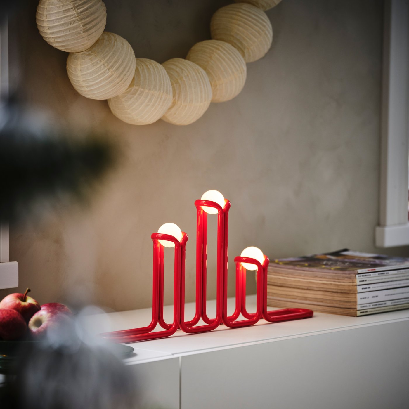 Three small round globe lights are held in place by red rounded decorative shapes. The three lights are in a line, at different heights, giving a modern candelabra impression.