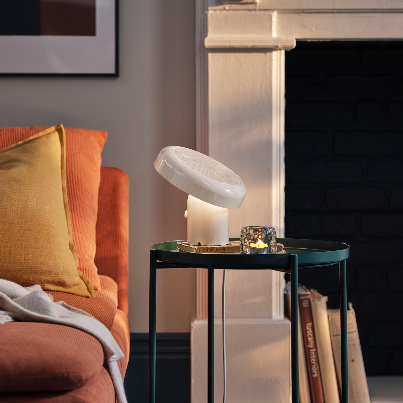 A white speaker lamp with downward light on black bedside table next to orange bed, with yellow pillow, white walls, and a fireplace in the background.