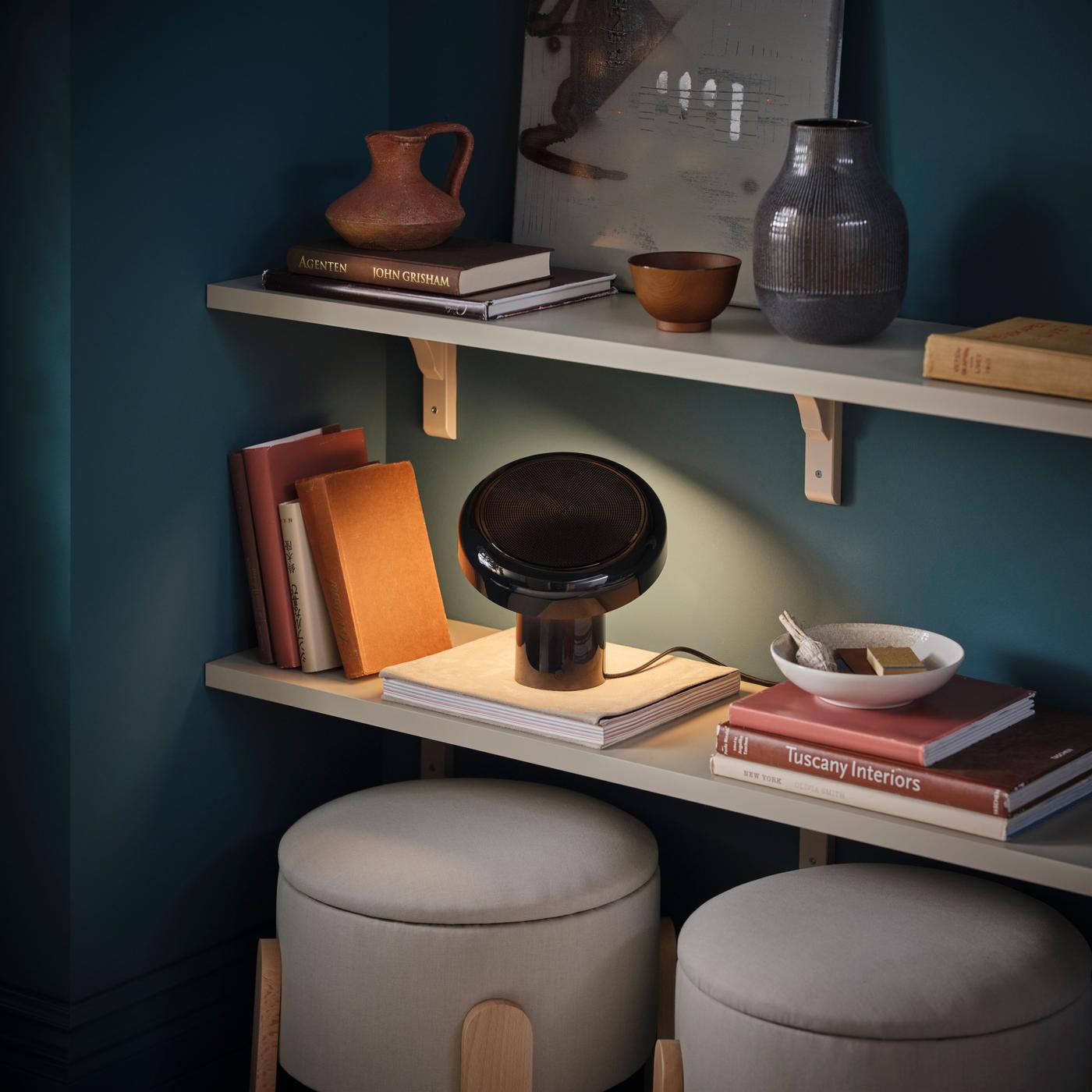 A black speaker lamp with a soft downward light, placed on a shelf filled with books and objects, with stools below.