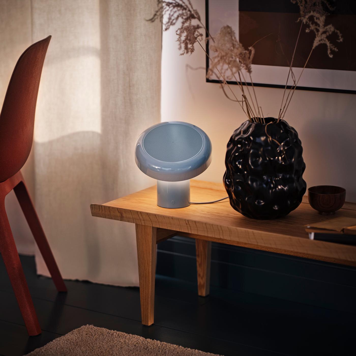 A grey-blue speaker lamp on a wooden desk, beside a black vase and softly lit white curtains.