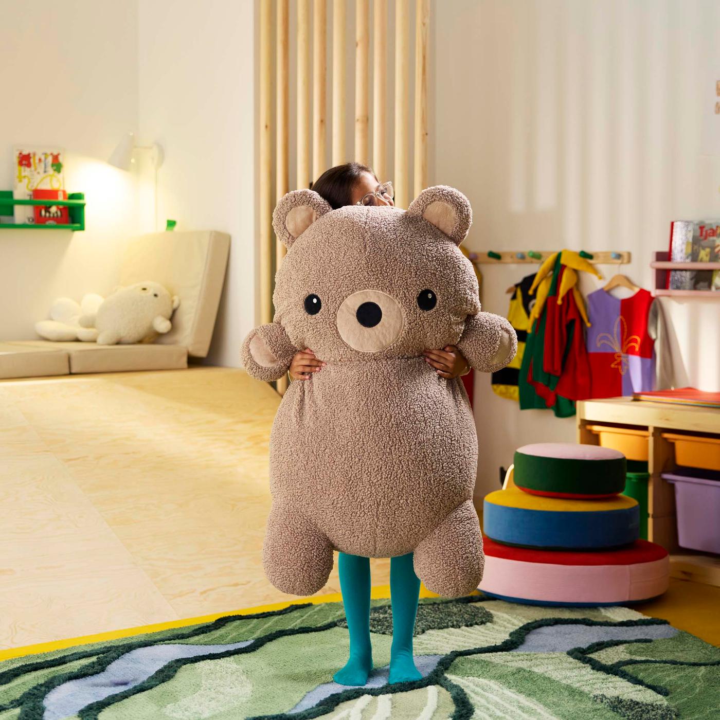 A child stand in a colourful living room hugging a large bear soft toy.