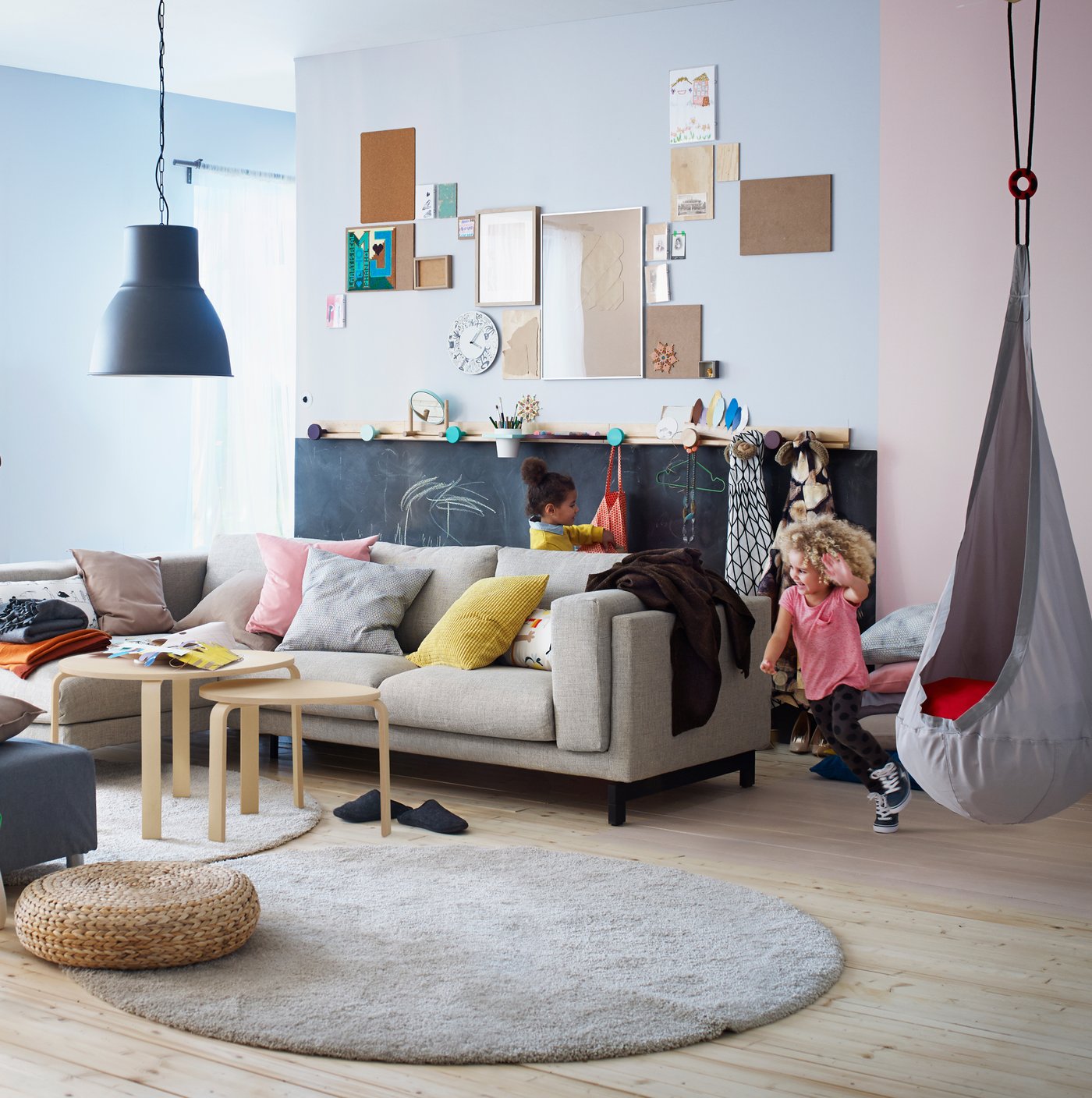 A child draws on a chalkboard surface in the back of a colourful, playful living room area, while another child runs around the sofa.