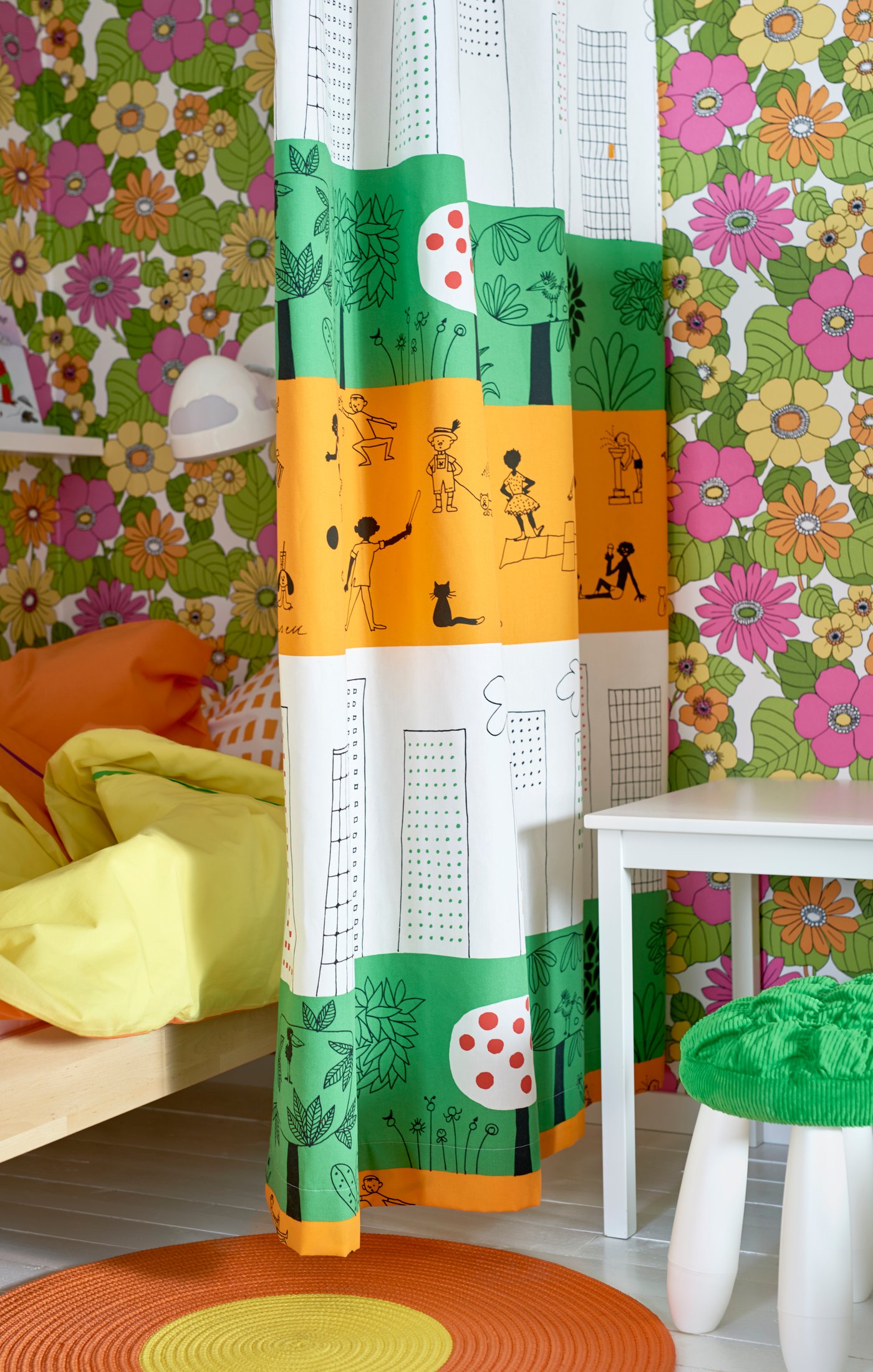 A green, white and orange striped curtain with graphic drawings, in front of a bright floral patterned wall and a small bed.