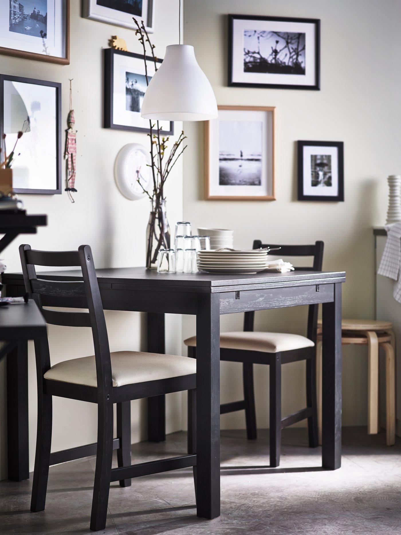 A small, dark wood dining table and two chairs are placed near a wall. On the wall is lots of framed photos and images. 