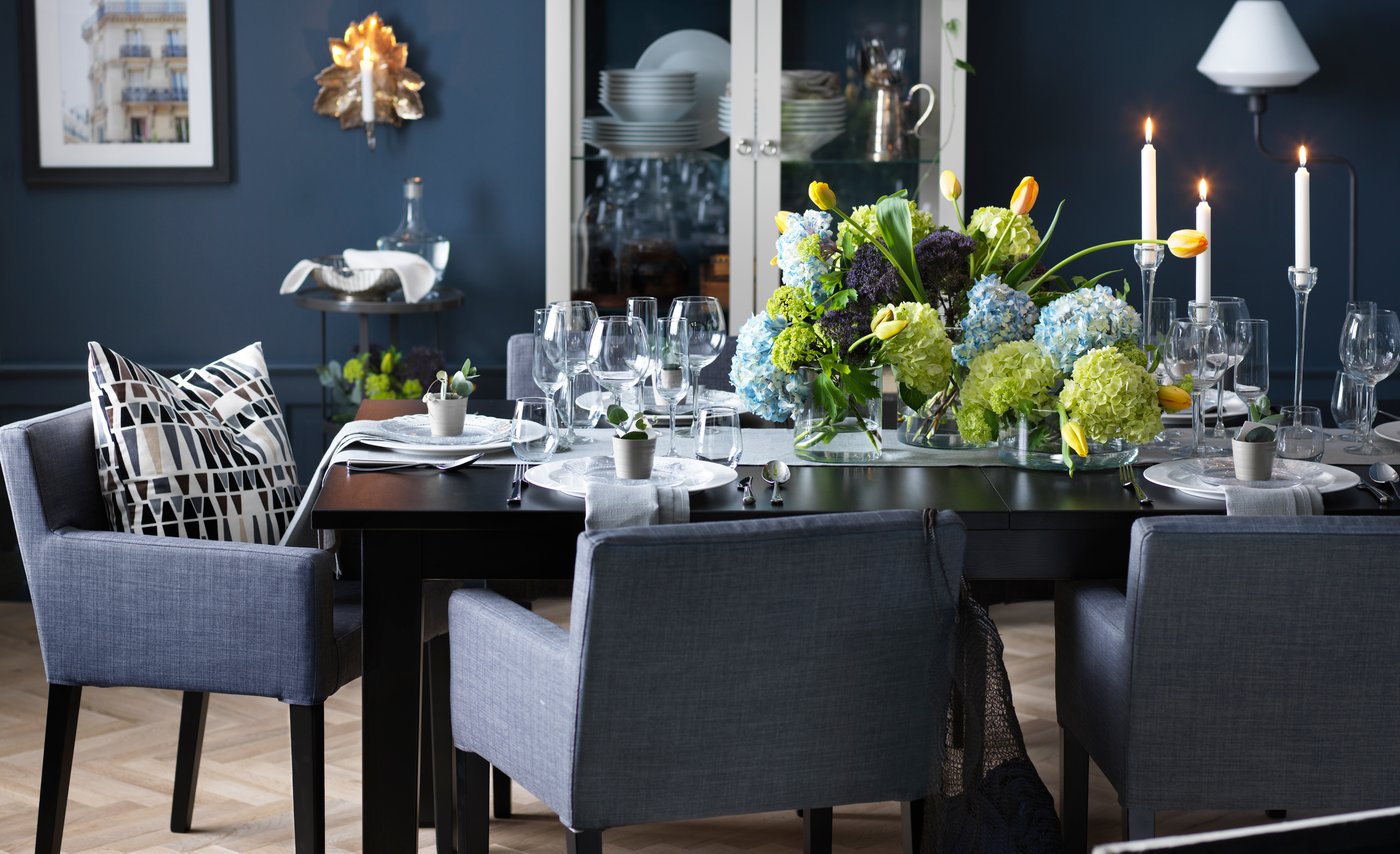 A blue dining room with a grey upholstered dining room set and flowers and candlesticks decorating the table.