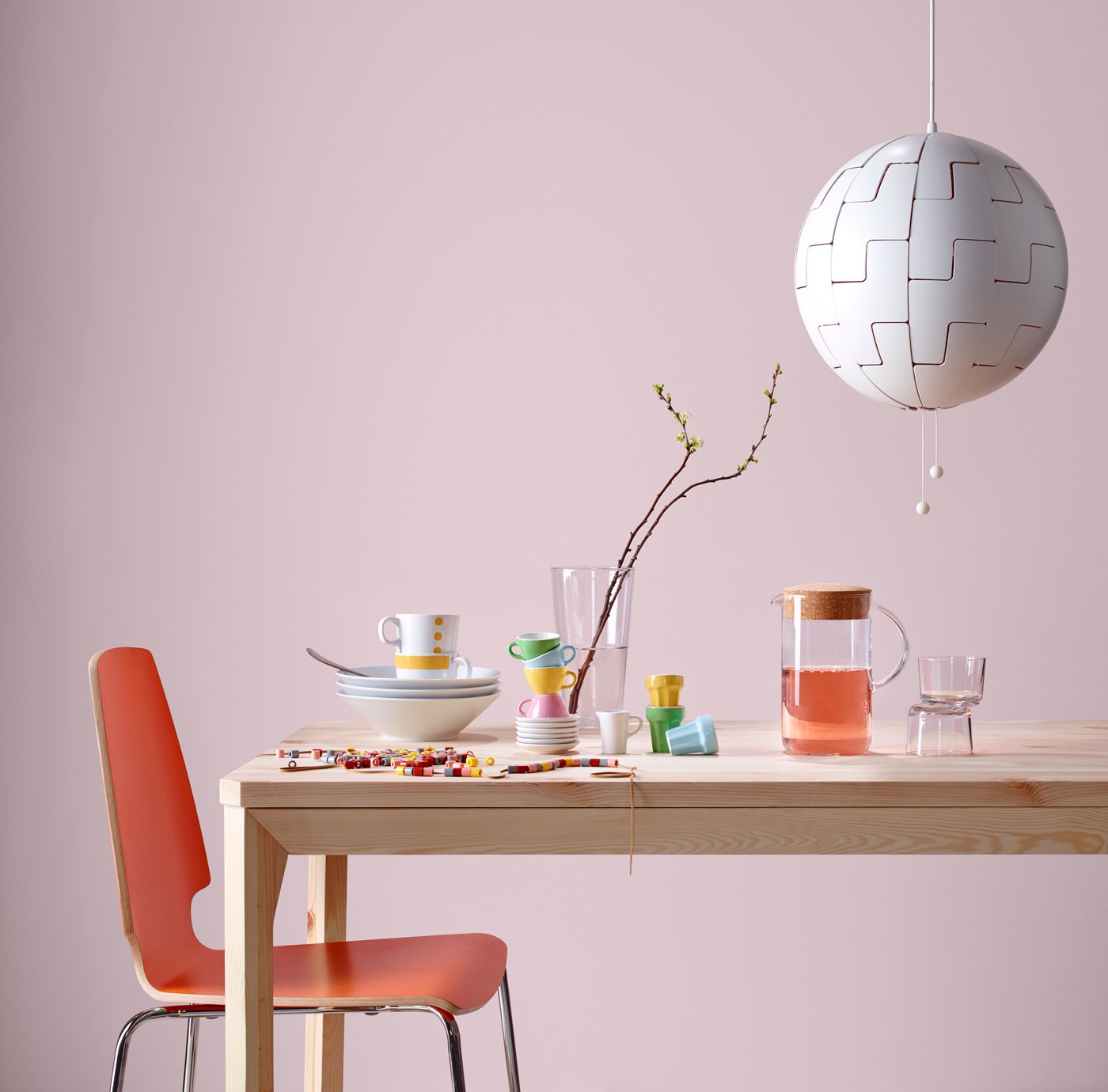 An orange chair with a wooden table propped with colourful plates and cups, against a pink background.