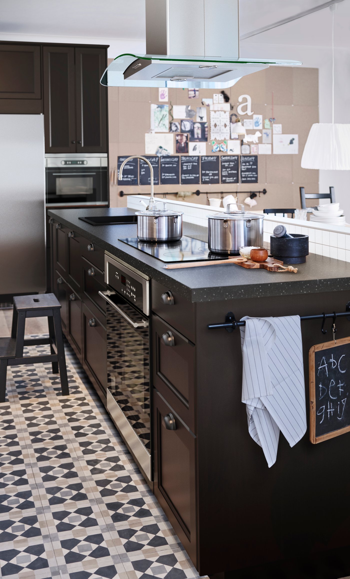 A brown kitchen island in a kitchen, with a large cork board on the wall, filled with chalk boards and papers.