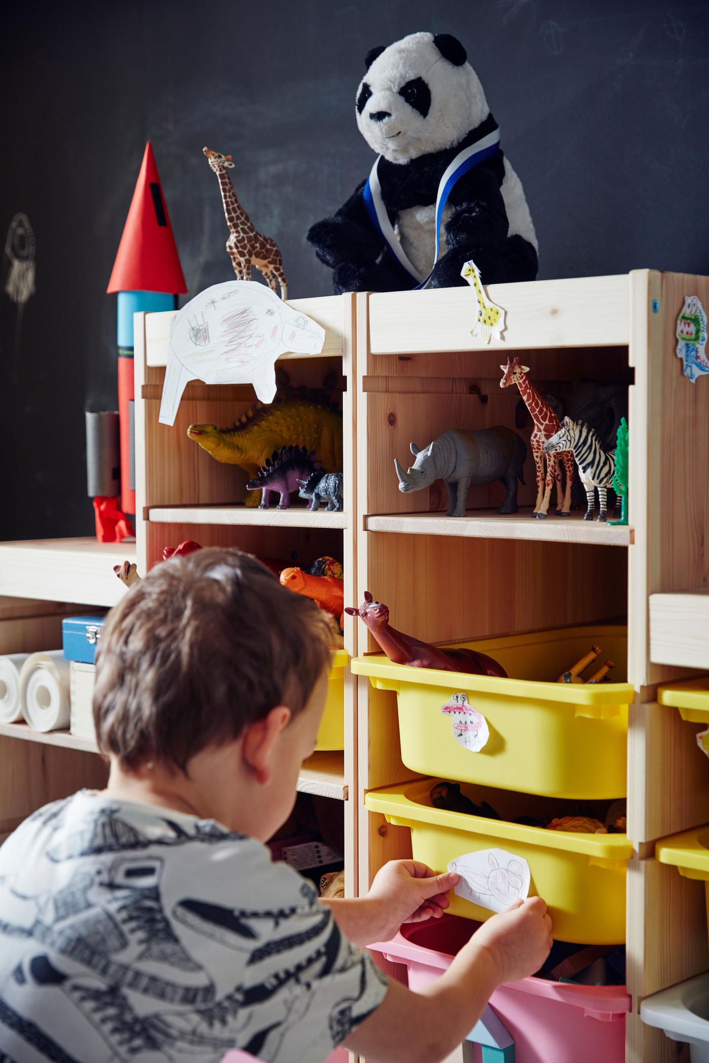 A boy standing in front of an IKEA store solution made of wood. On top several different toys and inside several wooden planks and two yellow plastic drawers oh which one he's holding.