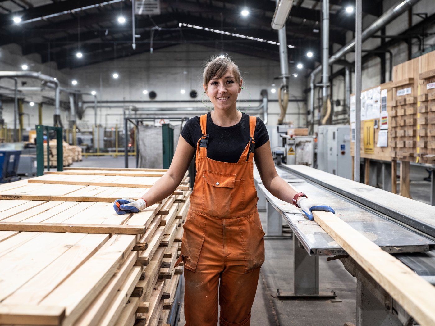 An IKEA co-worker in orange carpenter’s trousers and gloves in a factory, standing next to a large pile of wooden planks.