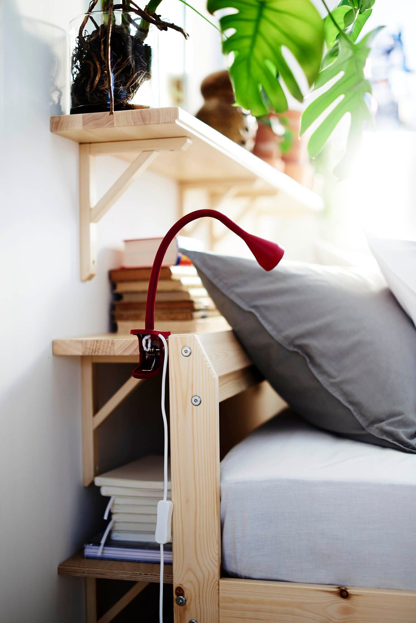 A red reading lamp clamped to a wooden shelf behind a bed. There is a shelf above covered in plants and one below with books.