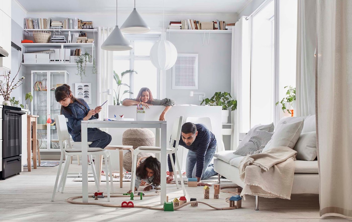 A family gather around a table in the living/dining room. A man kneels on the floor to play with a child, who is playing with a trainset.