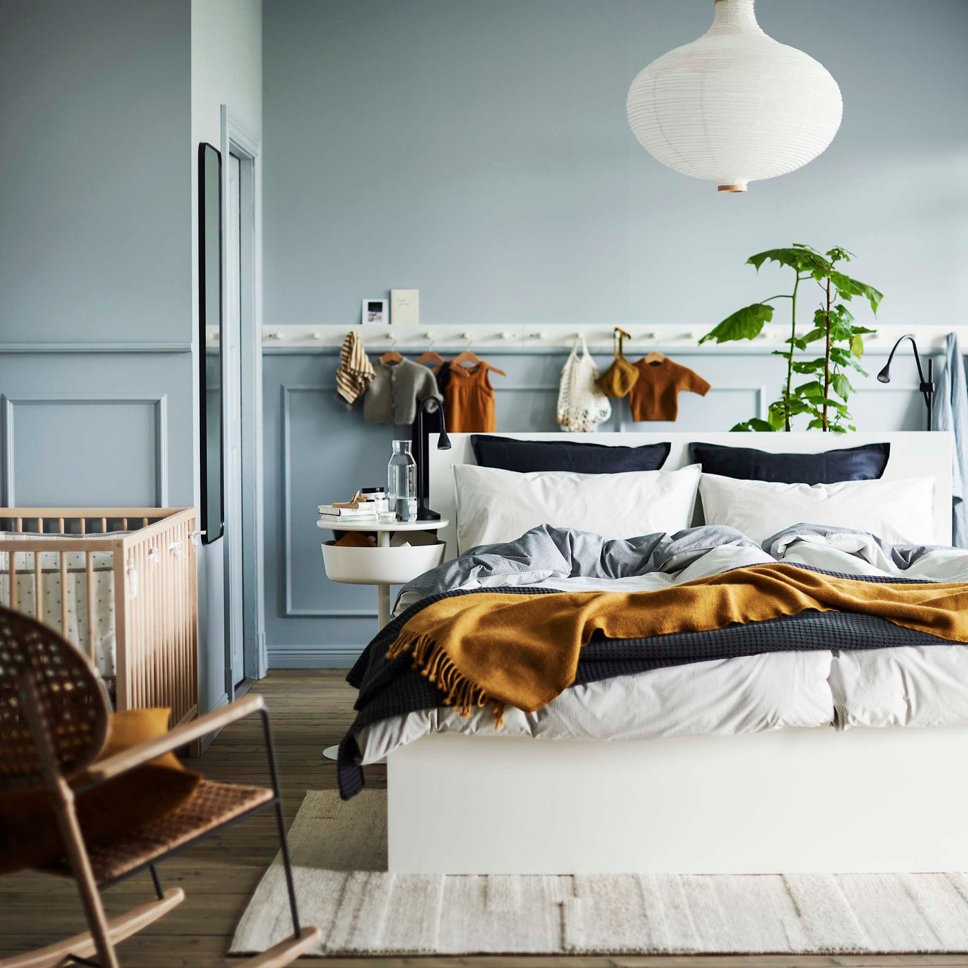 A blue bedroom with blue, grey and mustard bedlinen, in the 2021 IKEA catalogue. In the background hang baby clothes and above is a paper lampshade and a cot to the left.