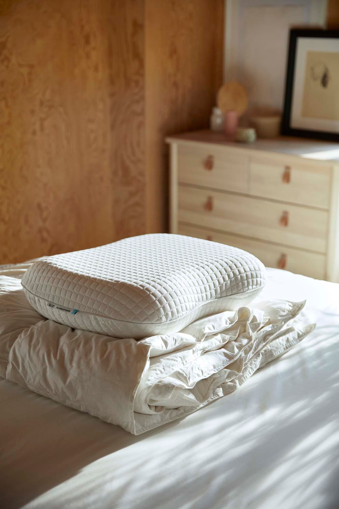 A KLUBBESPORRE ergonomic pillow and a folded duvet sit on top of a bed in a bedroom.
