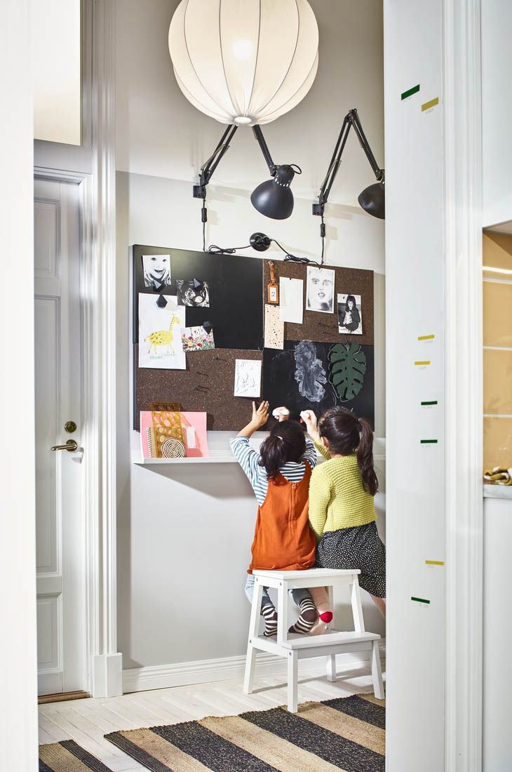 Two children stand on a white stool, they are sticking things on a notice board.