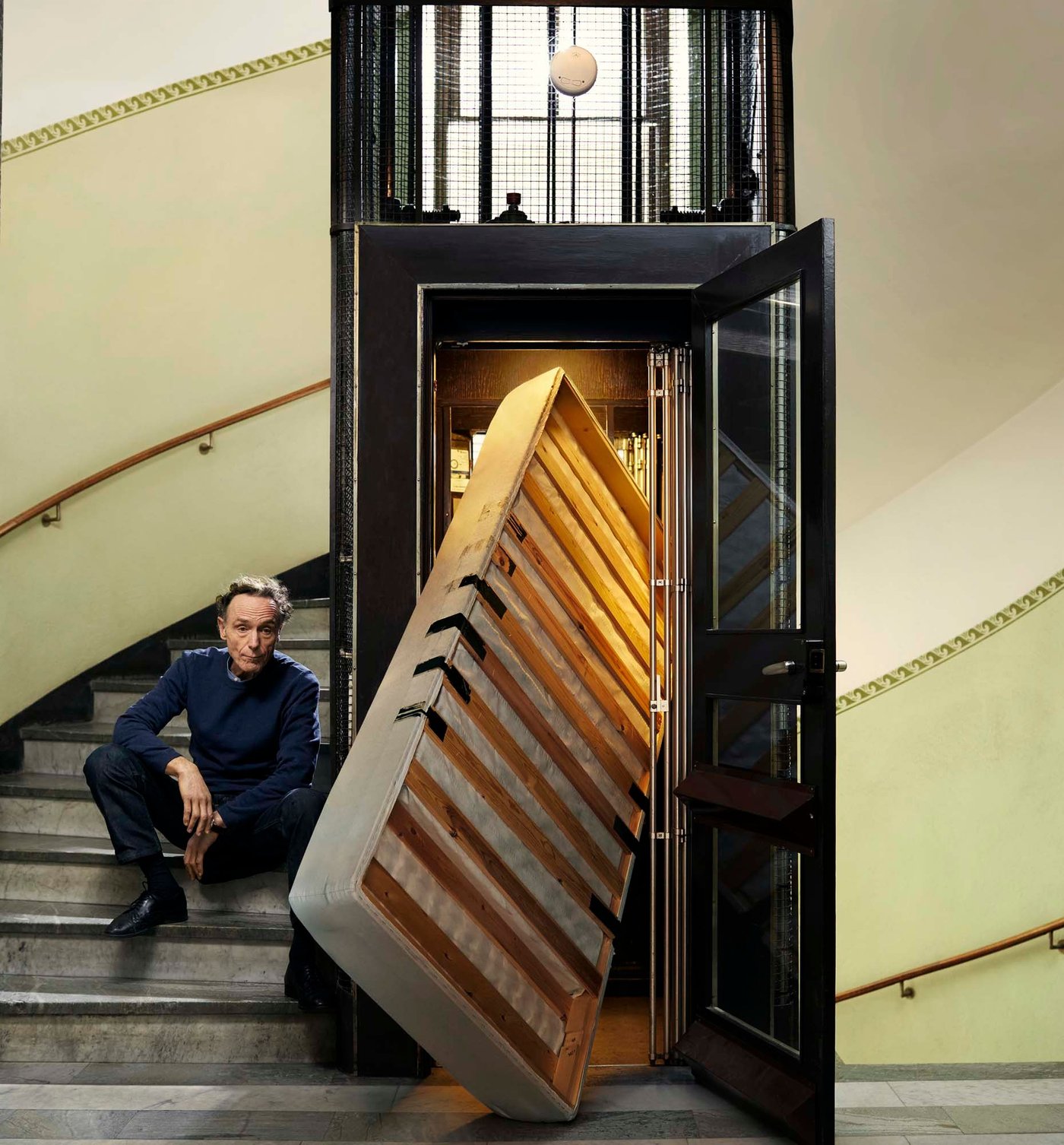 A person sits on a stairway in an apartment block. A mattress sticks out of an old fashioned lift – it doesn't fit.