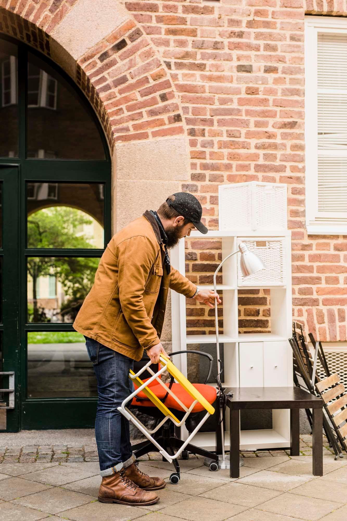 A person inspects a standing lamp and other items of furniture left on a pavement outside a brick building.