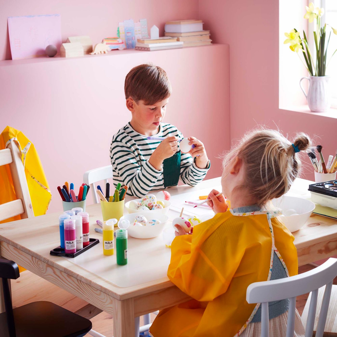 Two children crafting at a wooden table with a pale pink backdrop.