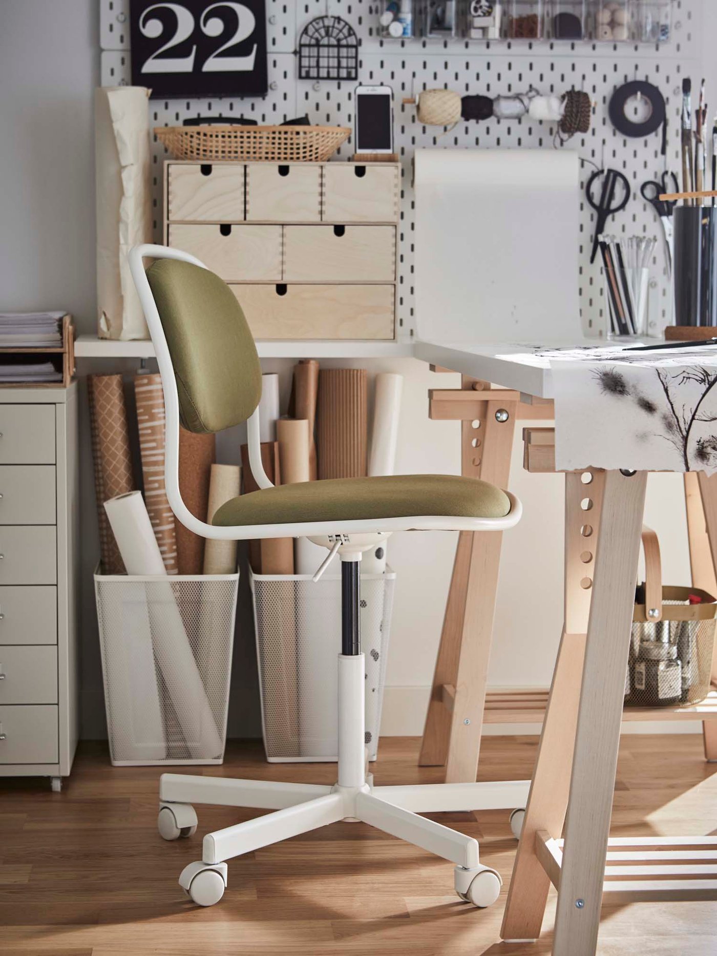 A green office chair sits in front of a desk with mesh baskets, wooden storage drawers and a wall organiser filled with stationery in a creative style workspace.