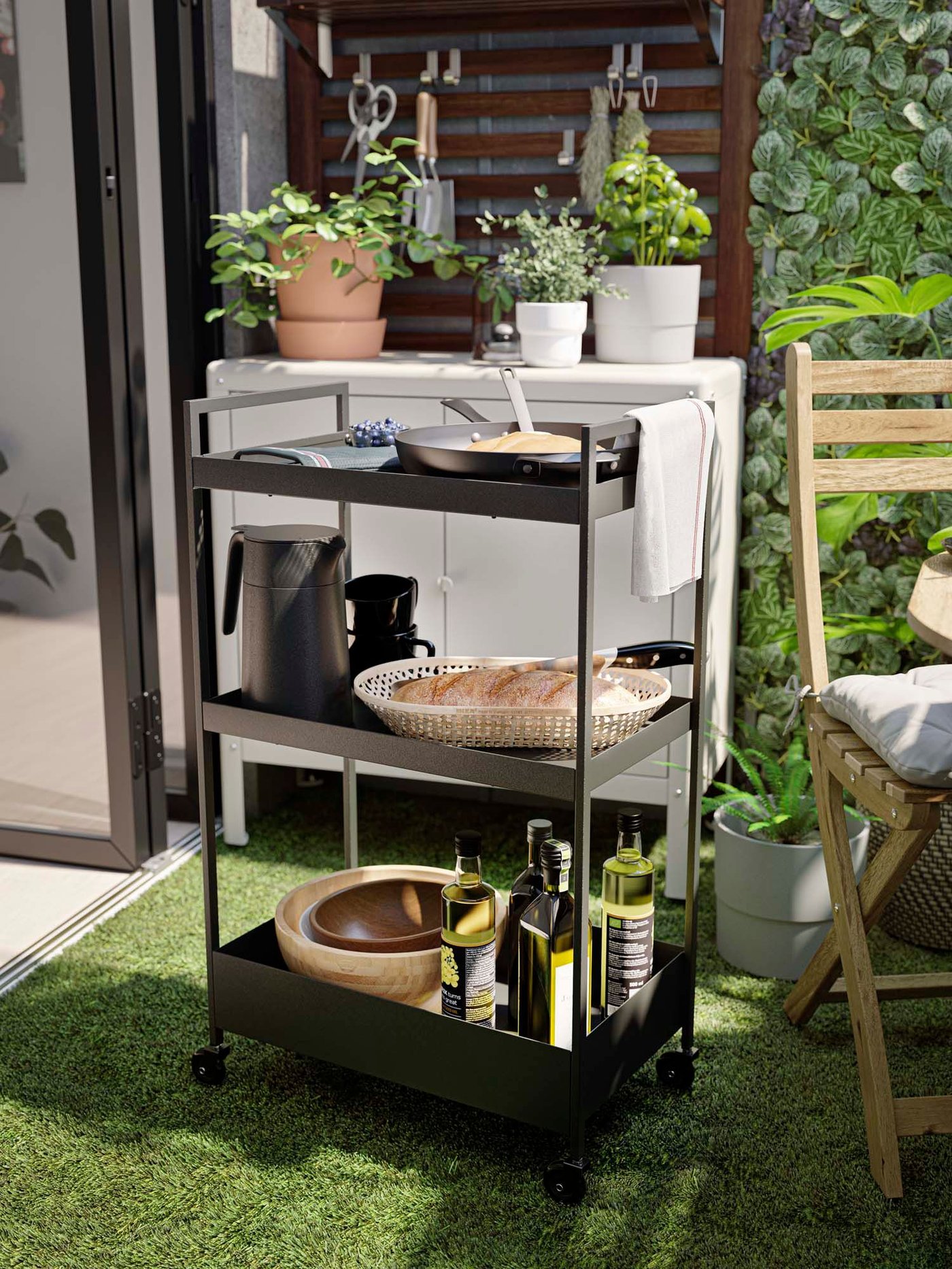 An outdoor area with a black NISSAFORS trolley carrying bowls, jugs, bottles of olive oil and bread in a basket.