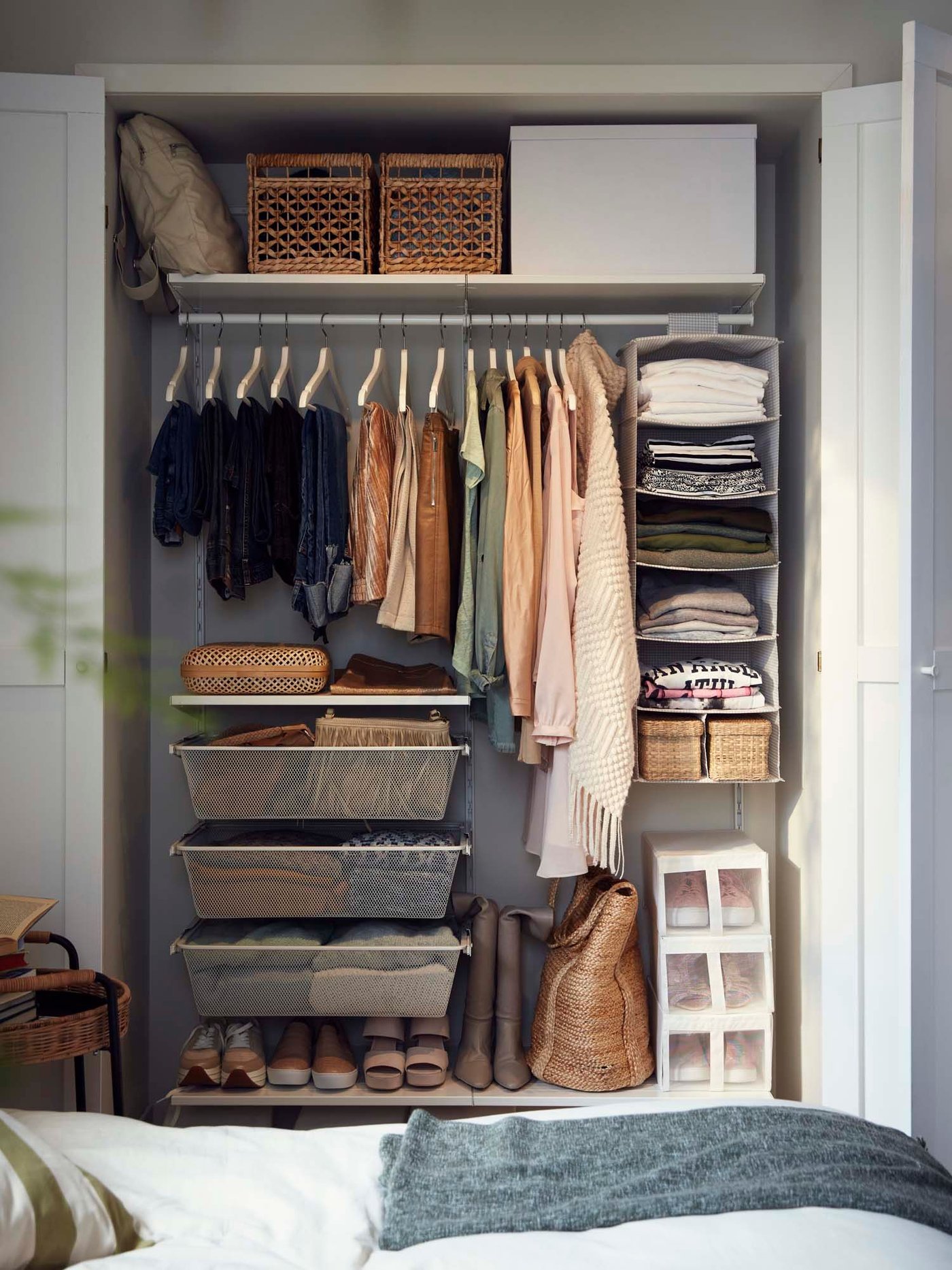 An open wardrobe in a bedroom reveals clothes on a rail, storage baskets below and shoes on bottom. In front of the wardrobe is a bed.