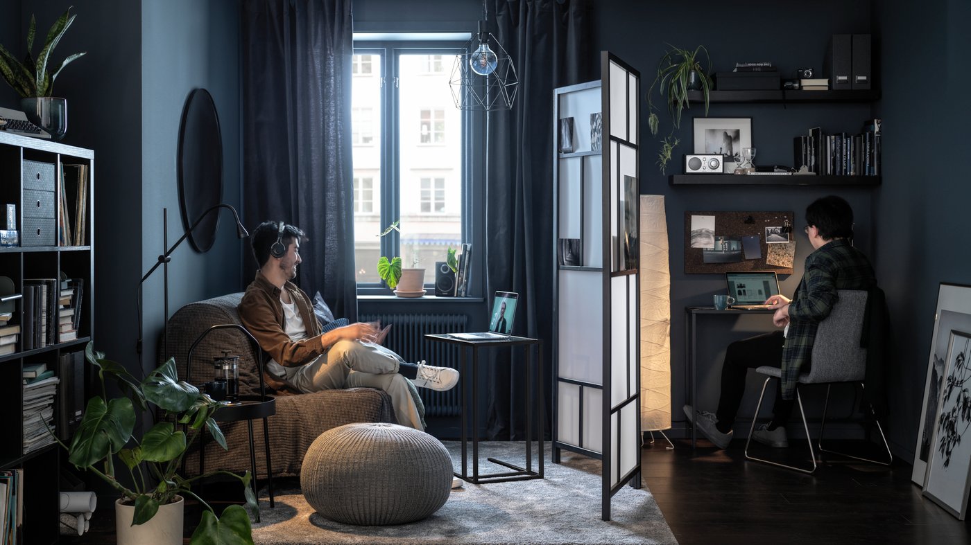 A room divider stands between a person working at a desk and a person on a video call in a dimly lit dark blue-grey living area.