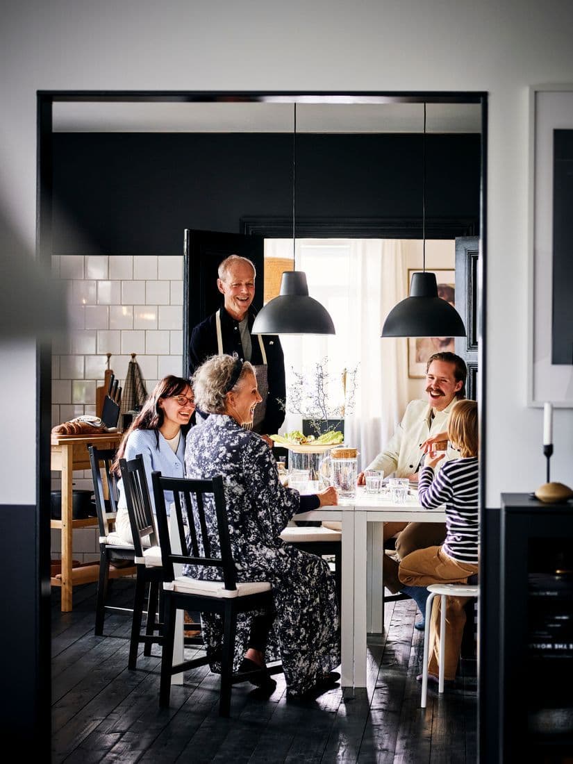 A family of five gathered around a white dining table in a dining area with black-painted hardwood floors and white-tiled walls.