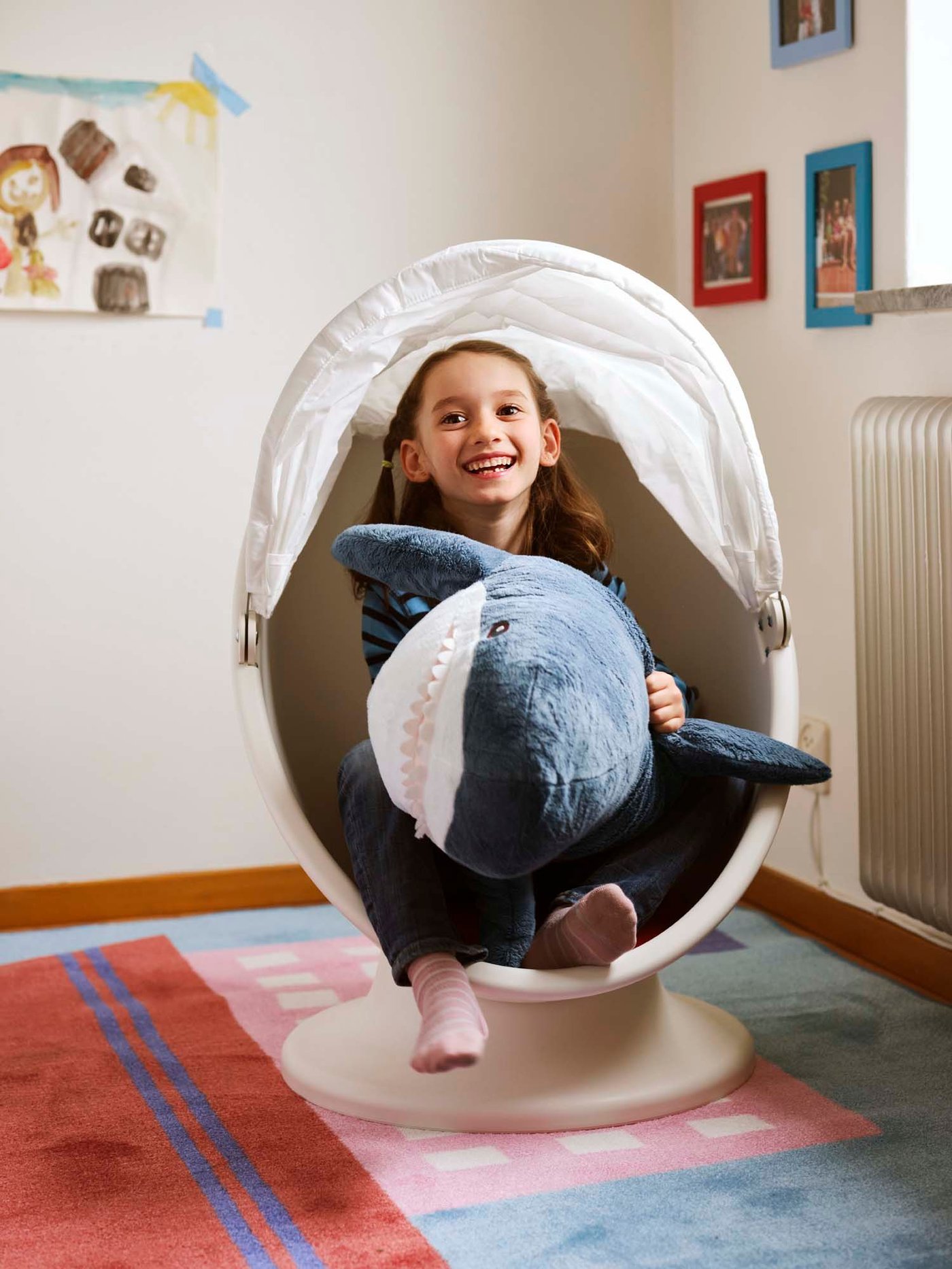 A girl sits in an egg-shaped chair in her bedroom. she is playing with a BLÅHAJ shark and smiling.
