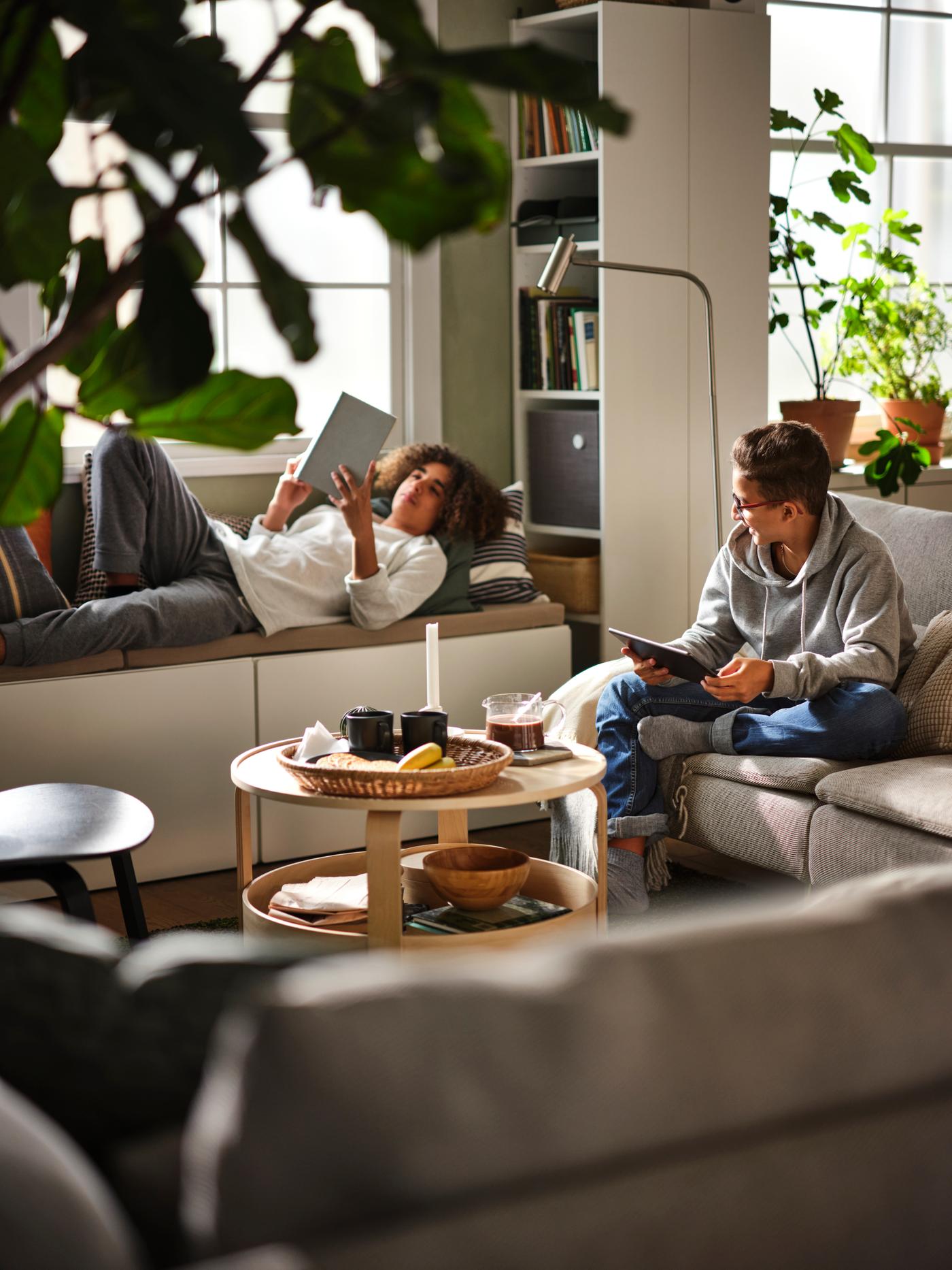 In a living room, a young person sits on a sofa looking at a tablet device, while another youth lays on a window seat, reading.