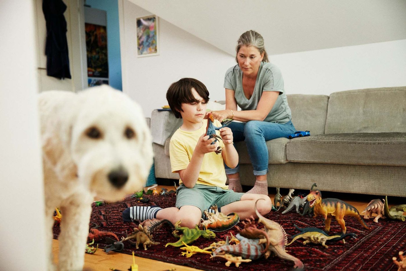 A woman sits on a sofa, watching her son play with his toys on a rug on the floor. In the foreground, a dog looks at the camera.
