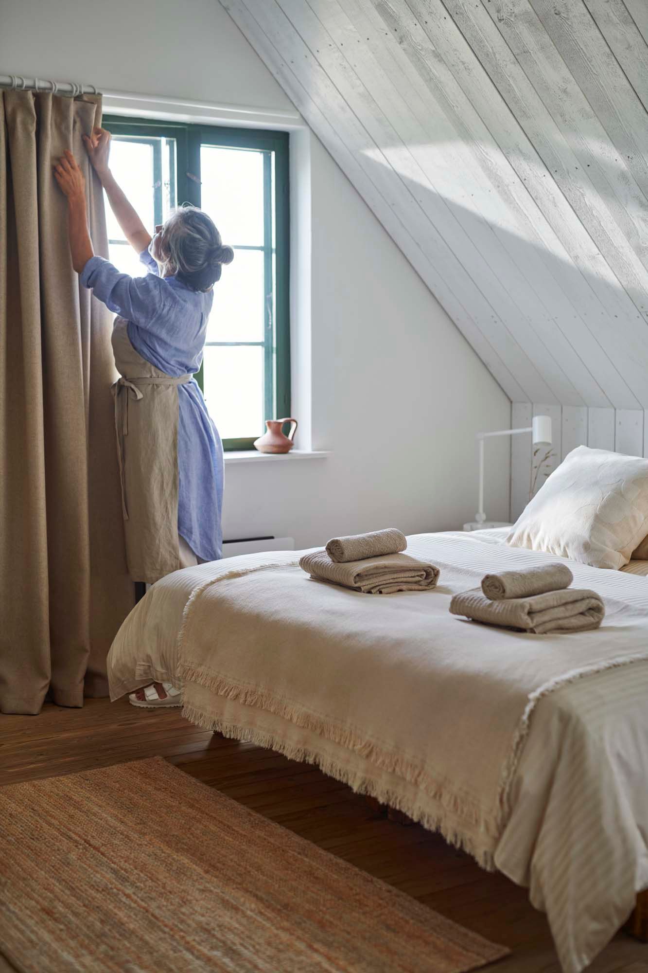 A woman stands in front of a bedroom window, next to a bed. She is fixing a pair of light coloured curtains.