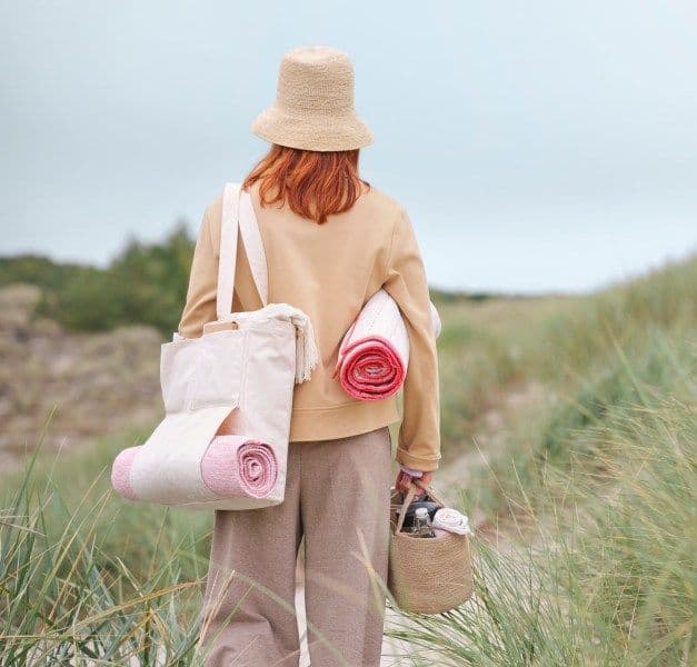 The back of a woman, dressed in a hat, carrying a bag on her left shoulder, walking away on the beach.