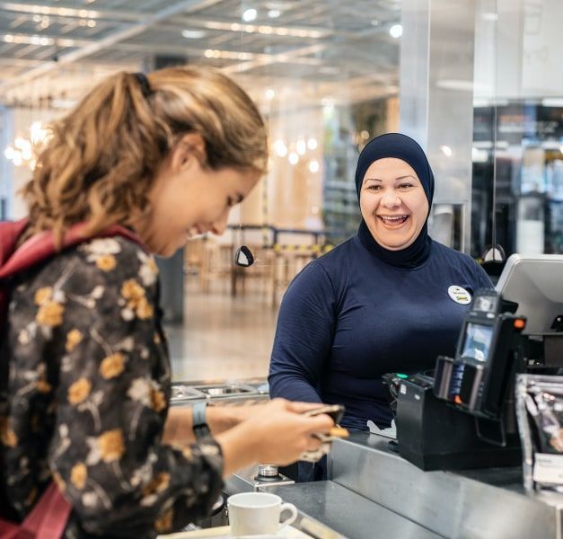 An IKEA restaurant co-worker at a cash register and a customer that is paying are laughing together.