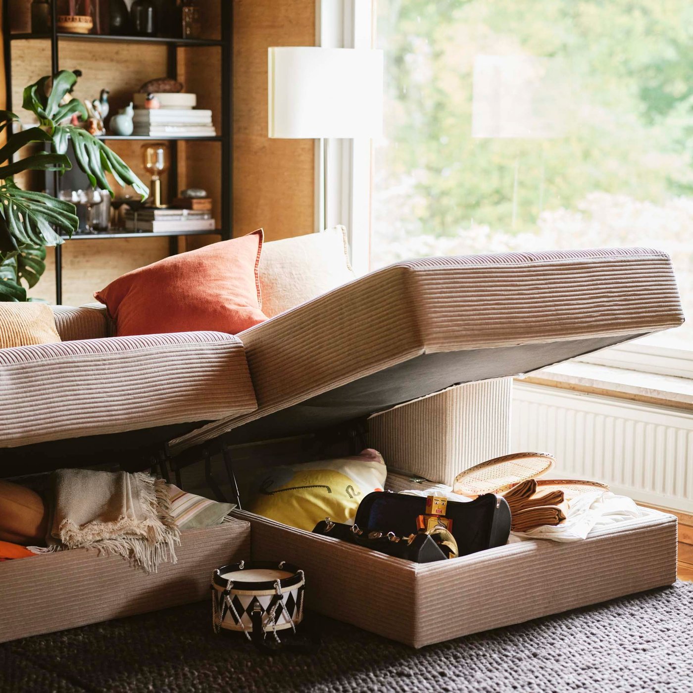 In a living room, a JÄTTEBO modular sofa is lifted, revealing its contents. In the background is a window with a verdant view.