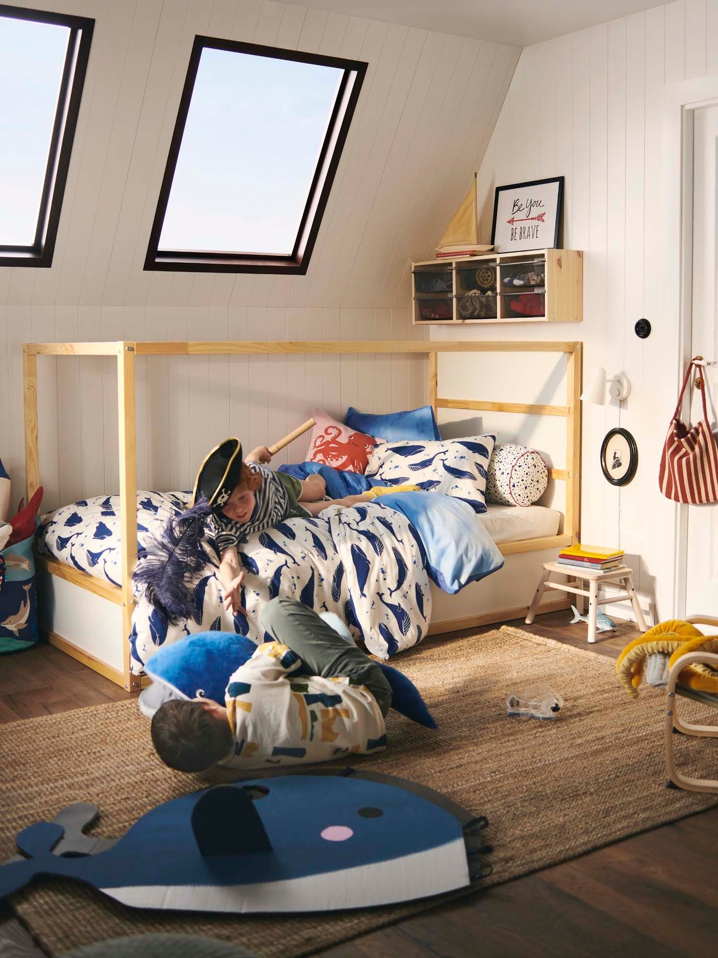 Two children play fight in a room. One is on the bed, wearing a pirate hat, while the other is on the floor with a toy shark.
