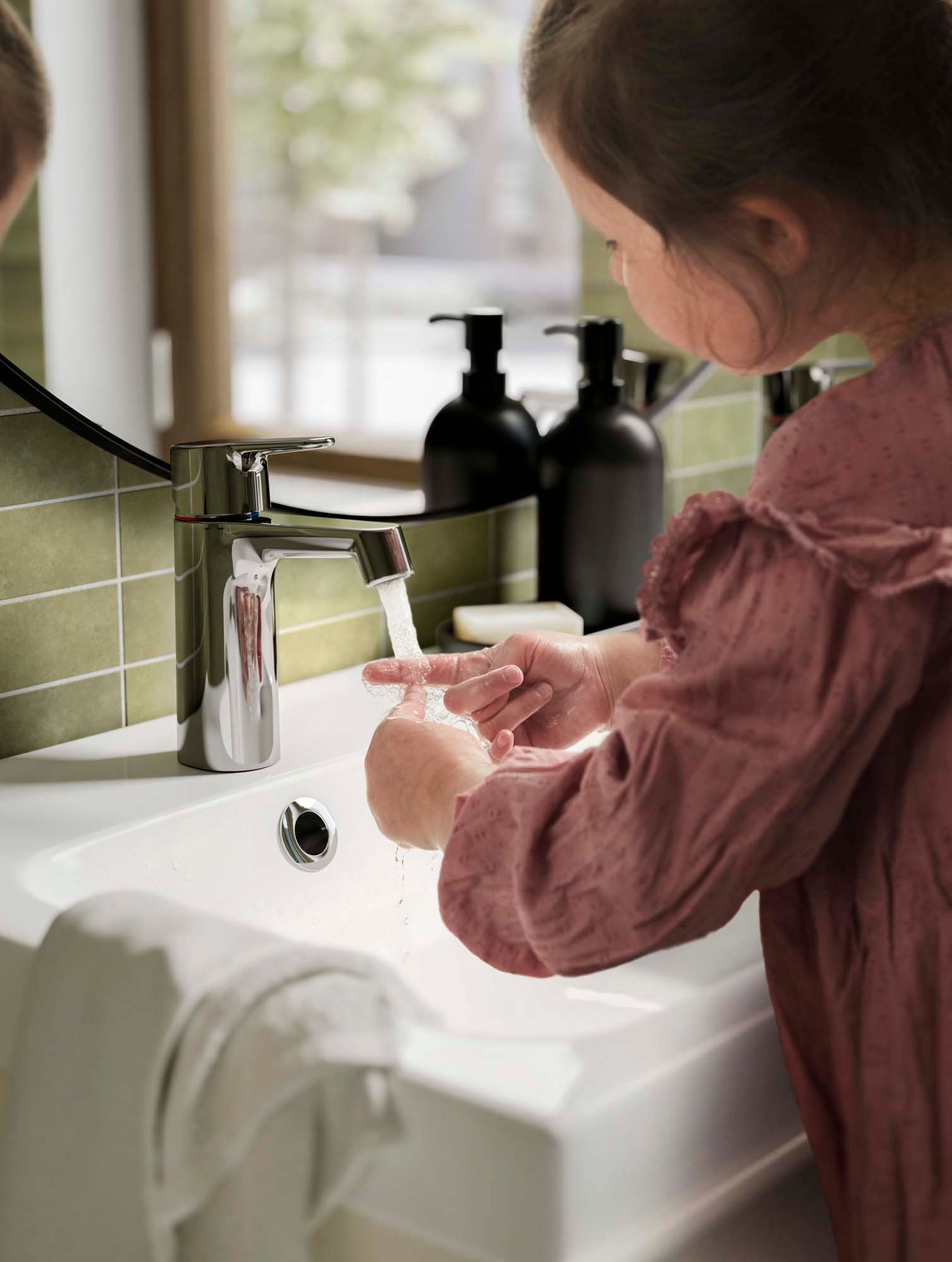 A child at a basin washing their hands under a running chrome-plated BROGRUND wash-basin mixer tap with strainer.