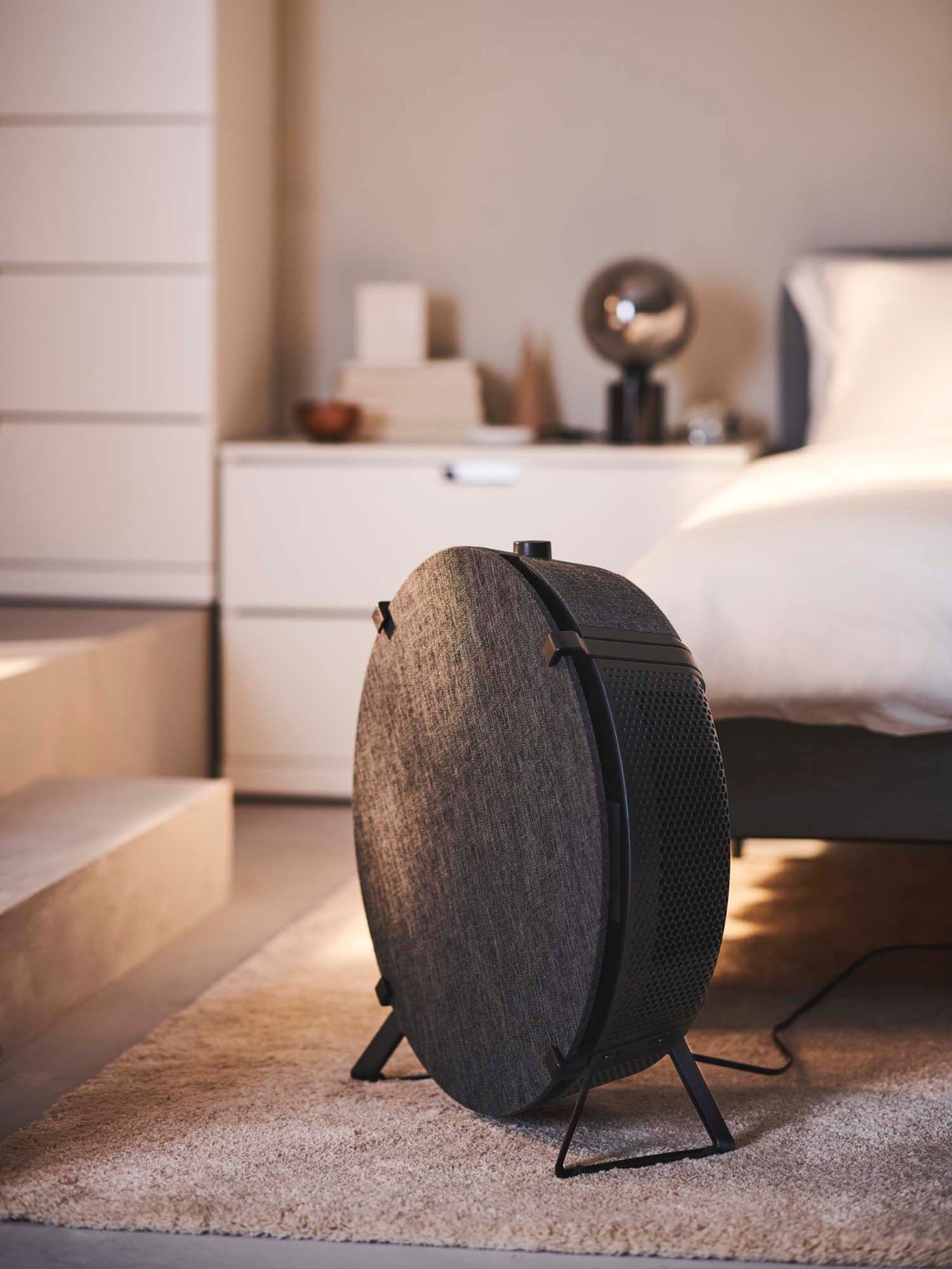 A bedroom with a black/smart STARKVIND air purifier on the floor at the foot of a bed.