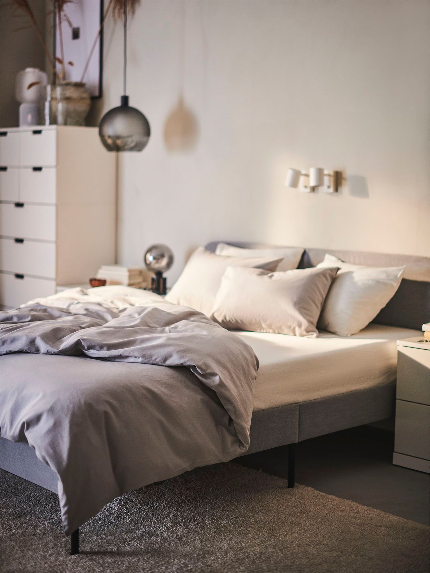 A dimly lit bedroom with a chest of drawers next to a light grey SLATTUM upholstered bed dressed in light grey NATTSVÄRMARE bedding.